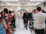 o	Bride and groom walking down the aisle holding hands with white carpet and floral archway, as guests applaud after Scottish last minute wedding