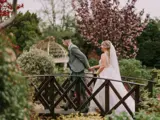 o	Bride and groom hold hands crossing garden bridge with lush greenery and blossom at The Lynnhurst wedding venue, after last-minute ceremony