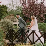 o	Bride and groom hold hands crossing garden bridge with lush greenery and blossom at The Lynnhurst wedding venue, after last-minute ceremony